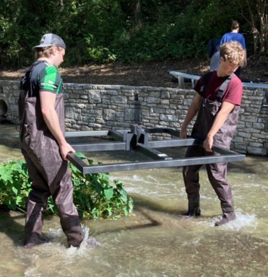 Two people in waders carrying a metal base frame through the creek